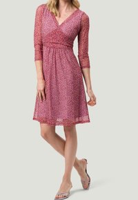 Robe rose à manches courtes avec un motif floral, décolleté en V et taille cintrée. Matière transparente, longueur genou, associée à des sandales à lanières.