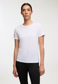 Donna con capelli scuri raccolti, che indossa una semplice camicia bianca a maniche corte e pantaloni neri, in piedi di fronte a uno sfondo chiaro.