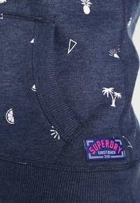 Felpa blu navy con stampe a tema tropicale bianche (ananas, triangoli) e polsini a coste, con un'etichetta del logo Superdry ben visibile.