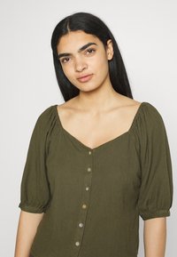 Top verde-azeitona com decote quadrado, mangas curtas em forma de balão e fecho frontal com botões coloridos como detalhes.