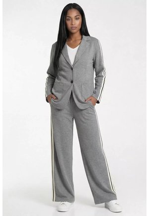 Kobieta w szarej marynarce na dwa guziki i pasujących szerokich spodniach z białymi bocznymi paskami, stojąca z rękami w kieszeniach.