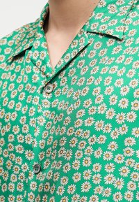 Πράσινο πουκάμισο με κουμπιά και λευκό και κίτρινο μοτίβο μαργαρίτας, φορεμένο με το γιακά ανοιχτό στο λαιμό.