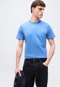 Νέος άνδρας με φωτεινό μπλε κοντομάνικο μπλουζάκι και μαύρο τζιν, κρατώντας ένα σκούρο μπουφάν στο αριστερό του χέρι, κοιτάζει προς τα πλάγια.