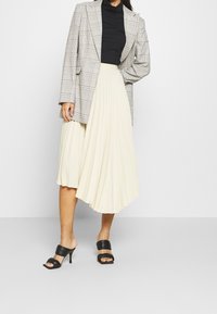 Blazer oversized a quadri grigi, dolcevita nera aderente e gonna midi plissettata color crema abbinate a mule nere con tacco.