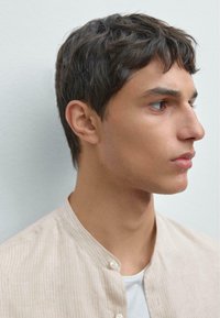 Profilansicht einer Person, die eine hellbeige, knöpfbare Jacke mit Stehkragen über einem weißen Hemd trägt. Kurze, strukturierte dunkle Haare.
