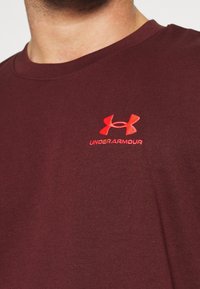 Vínová tričko s kulatým výstřihem vyrobené z měkké směsi bavlny, s červeným vyšitým logem Under Armour na levé části hrudi.