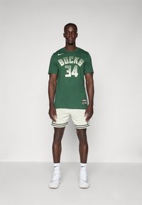Žalias Nike sportinis džersis su auksiniais užrašais "BUCKS 34", derinamas su baltais perforuotais šortais su juodu apvadu, dėvint baltus sportbačius.