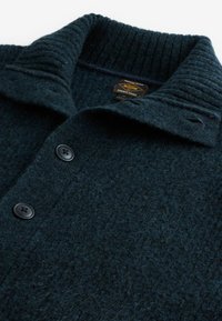 Pulover din lână de un albastru închis, cu guler ribbed, două nasturi negri și țesătură texturată. Eticheta este vizibilă cu inscripția "DURABLE GOODS".