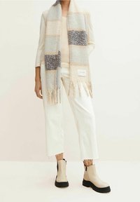 Beige- och grårandig scarf med fransar, bärs över en ljus tröja och vita capribyxor, ihop med ankelstövlar med svarta sulor.