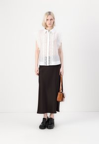 Blusa bianca trasparente con dettagli in pizzo e bordi a frange, abbinata a una lunga gonna marrone scuro. La modella tiene in mano una piccola borsa marrone e indossa scarpe con platform nere.