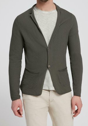 Hombre con blazer texturizado verde oscuro con dos bolsillos delanteros sobre camisa gris claro y pantalones beige contra fondo liso.