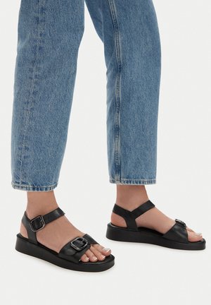 Sandale negre din piele cu o talpă groasă, dotate cu catarame ajustabile și o curea în jurul gleznei. Purtați cu blugi din denim albastru.