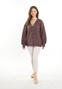 Barna cable-knit kardigán V-nyakkal, gombos elülső résszel és texturált puff ujjal. Beige felsőn viselve világos színű, testhezálló nadrággal.