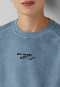 Lichtblauwe katoenen sweatshirt met een geribbelde halsline en zwarte bedrukte tekst, met de merknaam "SLVR STUDIOS" en een slogan eronder.