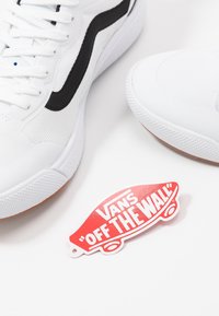 Weiße Sneaker mit schwarzem Streifen, Obermaterial aus strukturiertem Stoff und Gummisohle. Rotes Etikett mit "VANS OFF THE WALL" in fetter weißer Schrift.