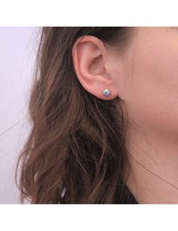 Orecchini a bottone rotondi con una gemma blu multifaccettata montata su una base in metallo argentato, indossati in un orecchio forato.