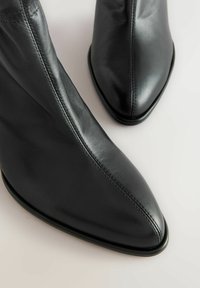 Kaksid sileda musta nahast teravatipulisi saapaid, mille keskmise õmbluse ääres on nähtavad õmblused, heledal pinnal.