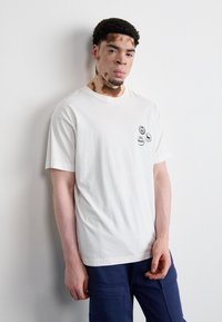 Biała bawełniana koszulka T-shirt z okrągłym dekoltem, z trzema małymi czarnymi naszywkami graficznymi. Noszona z granatowymi spodniami, na białym tle.