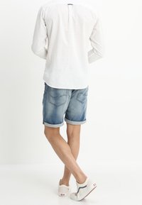 Camicia bianca a maniche lunghe con dettaglio sul collo sul retro, abbinata a pantaloni in denim blu. Scarpe casual con base bianca e accenti blu completano il look.