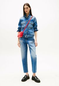 Chaqueta de denim con un degradado de azul claro a oscuro, combinada con jeans rectos de color azul. Complementada con un pequeño bolso cruzado rojo y mocasines negros.