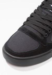 Nära bild av en svart sneakers tå med tyg- och mockadetaljer, svarta snören och en svart gummisula på en vit bakgrund.