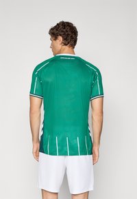 Käärus juustega mees, kes kannab rohelist spordisärki valgete triipudega ja valgeid lühikesi, seljaga vaataja poole ühevärvilise tausta ees.