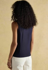 Marineblause ohne Ärmel mit einer figurschmeichelnden Passform, glatter Textur und Seitennähten. Kombiniert mit weißen Hosen mit Gesäßtaschen.