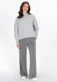 Grauer gestrickter Pullover mit hohem Kragen, kombiniert mit grauen, weiten Hosen. Der Pullover hat gerippte Bündchen und einen gerippten Saum. Das Model trägt weiße Sneaker.