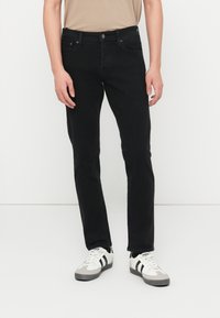 Άτομο που φορά στενά μαύρα τζιν, μπεζ μπλούζα και λευκά αθλητικά παπούτσια με μαύρες ρίγες, στέκεται μπροστά σε απλό λευκό φόντο.