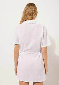 Vestido corto de lino blanco con cuello, mangas cortas y un cinturón atado a la cintura. Presenta una textura suave y un diseño sencillo.