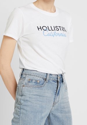 Persoon draagt een wit Hollister California T-shirt dat in een lichtblauwe, hooggetailleerde spijkerbroek is gestopt tegen een effen achtergrond.