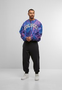 Übergroßer Pullover in Lila und Blau mit Muster und "COLUCCI" in weißen Buchstaben, kombiniert mit schwarzen Jogginghosen und weißen Sneakers.