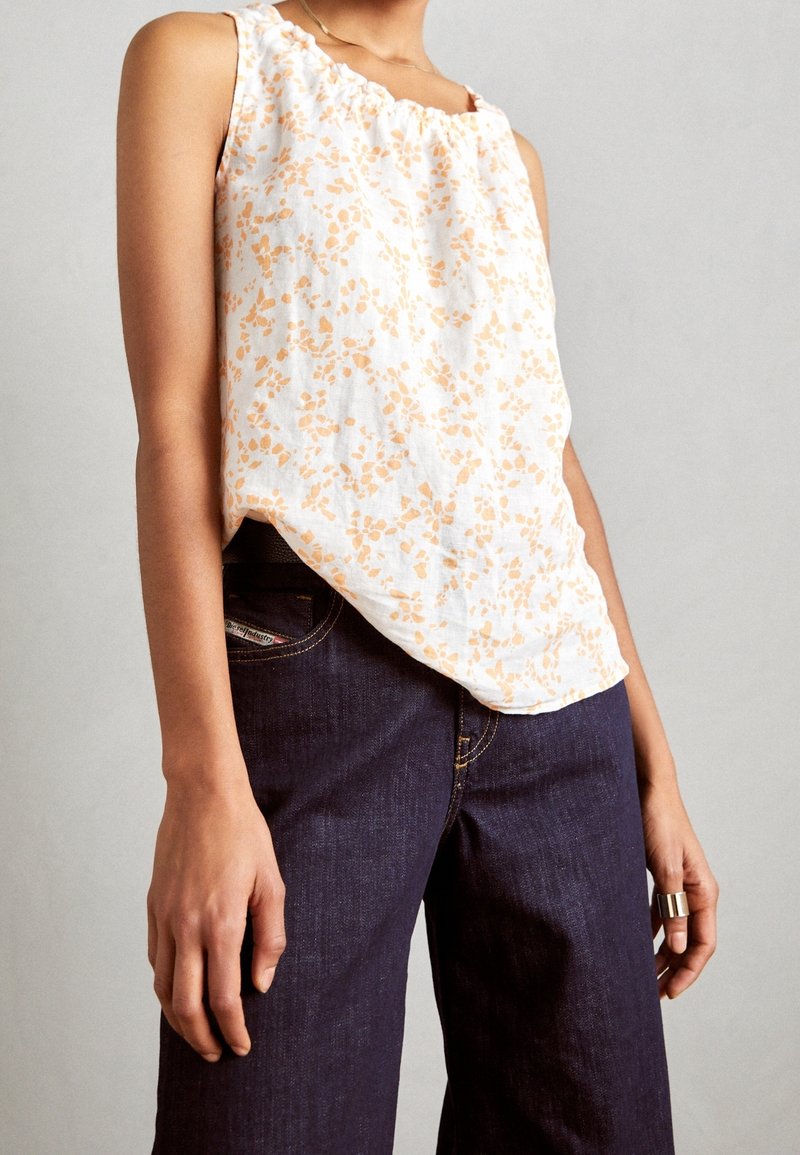 Débardeur blanc à motif floral orange, avec un col à volants et une silhouette ample. Associé à un jean large bleu foncé.