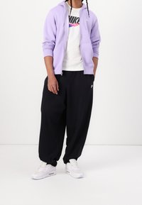 Fialová mikina na zip přes bílé tričko s černým logem Nike, doplněná volnými černými kalhotami a bílými teniskami.