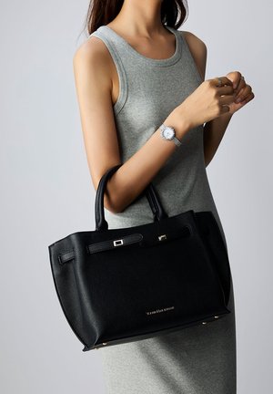 Black leather handbag with a textured surface, gold hardware accents, top handles, and a sleek design, held by a person in a gray tank top.