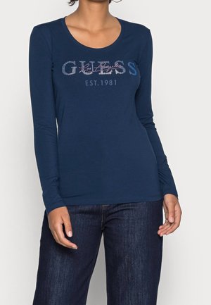 Nainen, joka pukeutunut tummansiniseen pitkähihamaiseen puseroon, jossa lukee "GUESS Est. 1981", ja tummansinisiin farkkuihin, yksiväristä vaaleaa taustaa vasten.