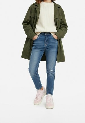 Persona vistiendo una chaqueta verde oliva, suéter crema, jeans azules y zapatillas rosa claro, con las manos en los bolsillos, de pie sobre un fondo blanco.