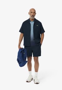 Kortärmad marinblå skjorta med knappar över en ljusblå polo, i kombination med marinblå shorts och vita sneakers. Jeanjacka i handen.