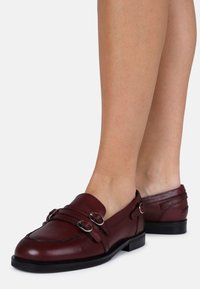 Mocassins em couro bordeaux com duplo fecho prateado, detalhes cosidos, bico arredondado e salto baixo em borracha preta.