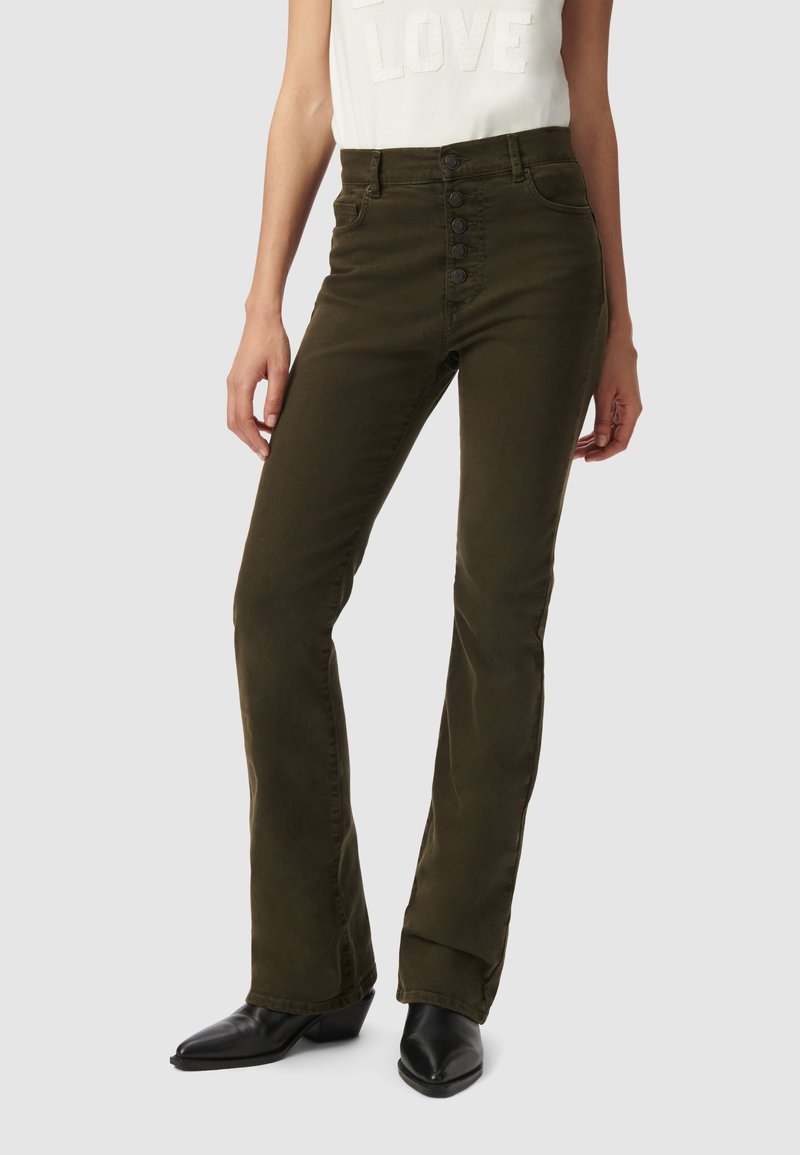 Vrouw in olijfgroene high-waist knopen-broek en zwarte puntige laarzen, met een witte "LOVE" top zichtbaar op de taille.