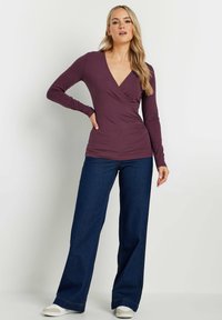 Langärmliges Oberteil mit tiefem V-Ausschnitt in tiefem Pflaumenrot, mit Wickeldesign. Kombiniert mit weiten, dunklen Jeans und weißen Slippern.