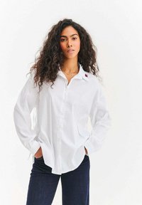 Biała koszula na guziki z kołnierzykiem i jednym czerwonym akcentem na lewym ramieniu, zestawiona z ciemnoniebieskimi dżinsami. Długie, luźne rękawy.