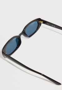 Μαύρα γυαλιά ηλίου με οβάλ πλαίσια, μπλε φακούς και χρυσές λεπτομέρειες στα βραχίονες. Γυαλιστερό φινίρισμα με λεία υφή.