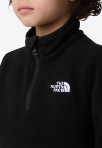 Pulover negru din fleece, cu fermoar până la jumătate și guler înalt, cu logo alb brodat pe pieptul stâng. Textură moale, fără modele vizibile.