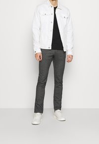 Giacca di jeans bianca, t-shirt nera, pantaloni grigi con texture e sneaker bianche indossati da una persona in piedi contro uno sfondo neutro.