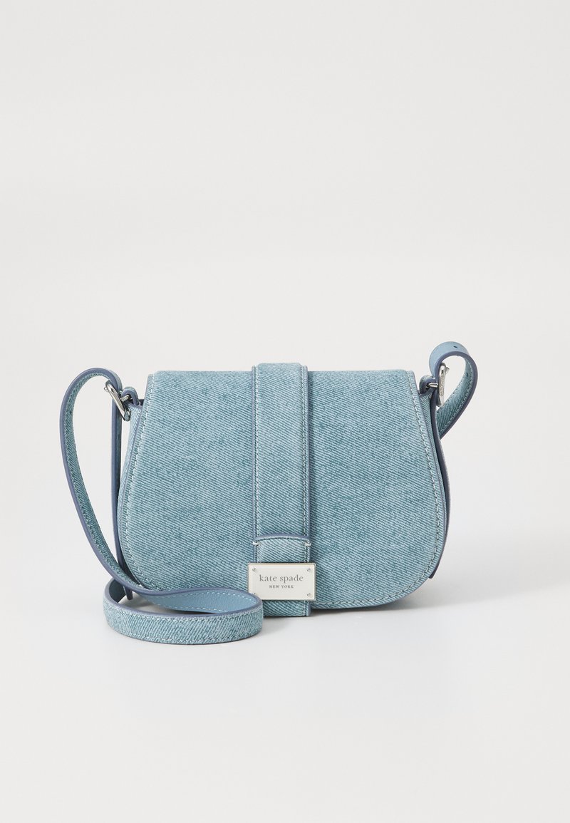 Umhängetasche aus hellblauem Denim mit glatter Textur, geschwungener Form, Klappe auf der Vorderseite und silberfarbenem Logo-Akzent. Abnehmbarer Gurt.