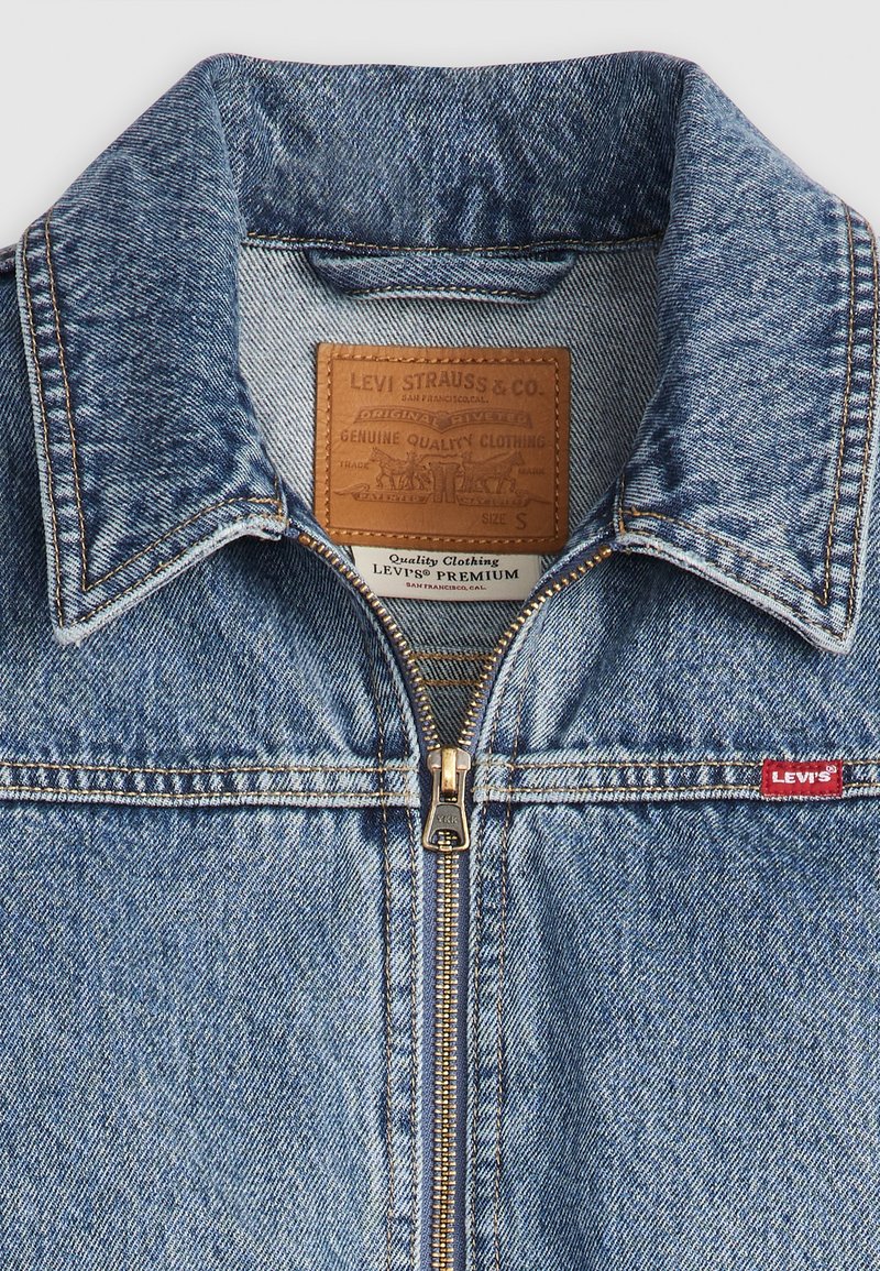 Chaqueta de denim en azul claro, con cuello, cremallera dorada, etiqueta de cuero y parche de logo rojo de Levi's. Textura suave con patrón desgastado.