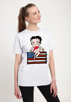 Hvid bomulds t-shirt med en tegneseriefigur med sort hår, rød top og guld accenter, siddende på et design af det amerikanske flag.