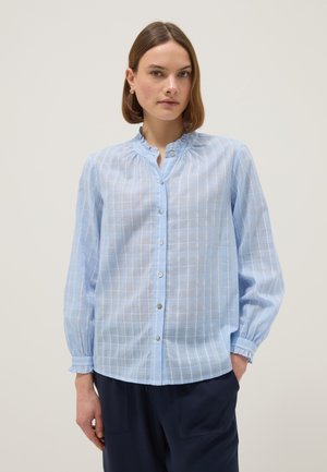 Femme portant un chemisier bleu clair à manches longues, boutonné sur le devant avec un motif à carreaux subtil et un col à volants, associé à un pantalon bleu marine.