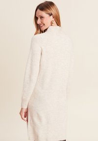 Robe pull beige clair avec un col montant, des poignets tricotés côtelés et des fentes sur les côtés. Texture douce avec un motif subtil, atteignant le mi-thigh.
